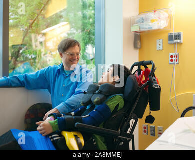 Smiling père assis à côté de fils handicapé en fauteuil roulant par lit d'hôpital, parler ensemble Banque D'Images