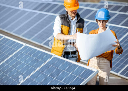 Deux ingénieurs ou d'architectes d'examiner la construction d'une centrale solaire, debout avec les bleus entre les rangées de panneaux solaires Banque D'Images