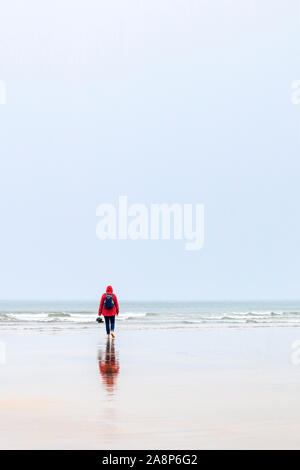 Une silhouette solitaire dans un anorak rouge à marcher vers la mer sur une plage balayée par des pluies et, convient pour une couverture de livre, Westward Ho !, Devon, UK Banque D'Images