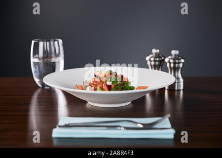 Focus sélectif de délicieux restaurant salade avec fromage servi en plaque blanche sur table en bois avec de l'eau, de couverts et de salières et poivrières sur bla Banque D'Images