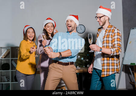 Happy businessman in santa hat holding partie cracker près de collaborateurs multiculturels avec verres de champagne Banque D'Images
