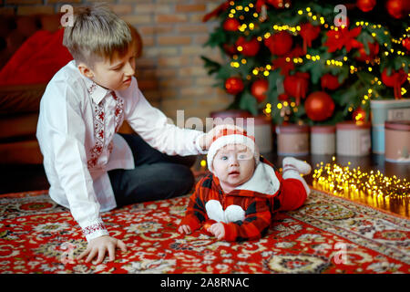 Frère avec petite sœur habillé en père Noël. au Nouvel An des arbres. Famille célèbre Noël. 6 mois petit garçon apprend à ramper sur backg Banque D'Images