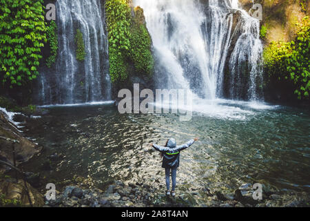 Young happy woman enjoying cascade dans Bali. Le port de l'imperméable gris de l'eau pulvérisée. Voyage en Indonésie. Banque D'Images