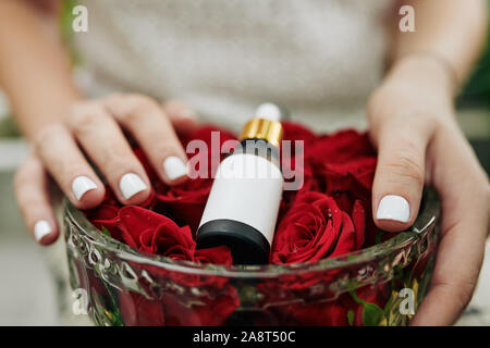 Mains de femme tenant un bol de cristal avec des boutons de fleurs et une bouteille d'huile de rose sur elle Banque D'Images