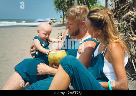 Drôle de famille heureux hipster pique-nique sur plage - mère, père nourrir bébé garçon. Petit-fils de manger des fruits avec plaisir, boire de noix de coco fraîche. Mode de vie sain, l'act Banque D'Images