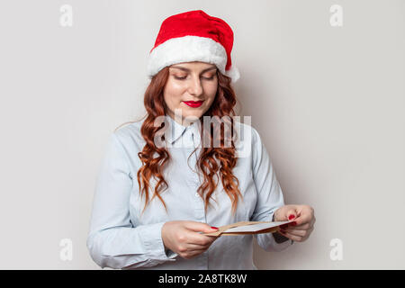 Jeune fille rousse dans un rouge santa hat lit une lettre, debout contre un mur gris dans le bureau. La préparation pour Noël et Nouvel An Banque D'Images