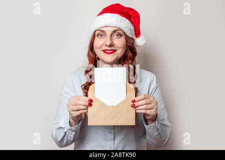 Young happy woman in santa hat et avec le rouge à lèvres rouge détient une lettre ou une liste souhaite avec une enveloppe de l'artisanat dans ses mains. Voeux de Noël et de rêves con Banque D'Images