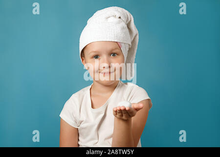 Petite fille avec une serviette blanche sur sa tête se lave le visage sur un fond bleu. L'enfant a de la mousse sur sa main. Concept d'hygiène quotidienne. Banque D'Images