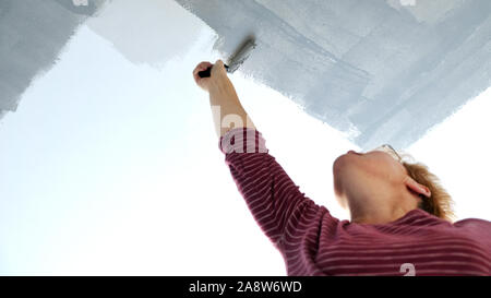 Une femme est la décoration de sa maison, à l'aide d'un rouleau de peinture à appliquer sur une peinture gris nouveau plafond blanc Banque D'Images