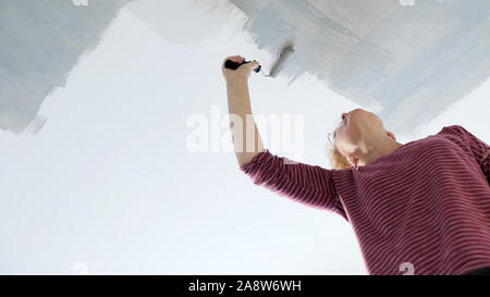 Une femme est la décoration de sa maison, à l'aide d'un rouleau de peinture à appliquer sur une peinture gris nouveau plafond blanc Banque D'Images