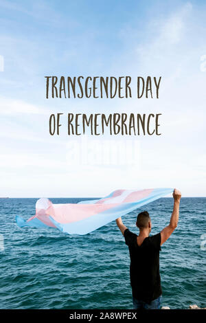 Une jeune personne de race blanche, vu de derrière, en face de la mer, tenant un transgender pride drapeau sur l'air et le texte remembranc transgenre de jour Banque D'Images