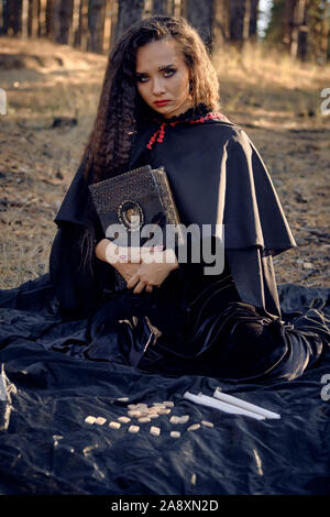 Jolie, méchants, enchanteresse aux cheveux longs dans une robe noire, avec le cap et le capot. Elle tient un livre, assis sur une couverture sombre avec les runes et ca Banque D'Images