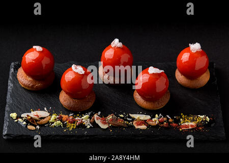 Mini tartelettes de fruits rouges sur le dessus vitré joliment disposés sur une plaque noire. Tartelette dessert festive moderne rouge et mousse dans le glaçage miroir shap Banque D'Images