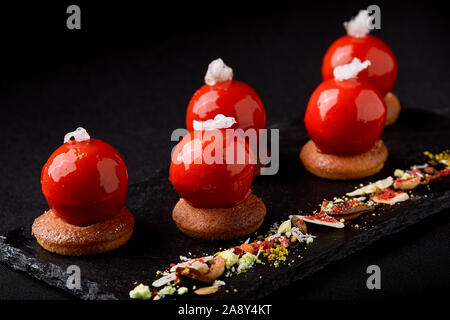 Mini tartelettes de fruits rouges sur le dessus vitré joliment disposés sur une plaque noire. Tartelette dessert festive moderne rouge et mousse dans le glaçage miroir shap Banque D'Images