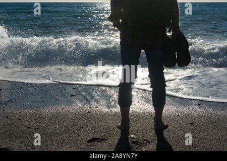 Pieds nus, un homme portant un petit sac à dos sur une plage tropicale avec ses chaussures dans ses mains, rétro-éclairé et à la recherche dans le soleil Banque D'Images