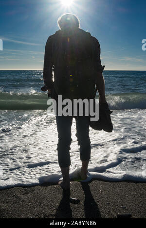 Pieds nus, un homme portant un petit sac à dos sur une plage tropicale avec ses chaussures dans ses mains, rétro-éclairé et à la recherche dans le soleil Banque D'Images