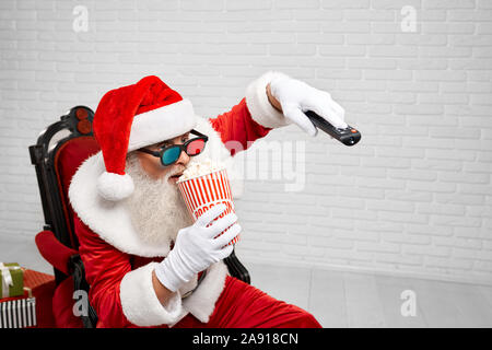 Heureux Père Noël authentique en 3D sans lunettes, assis sur chaise confortable en regardant la télévision et en mangeant du pop-corn savoureux de godet à rayures. Homme mûr se détendre après une dure journée de travail Banque D'Images