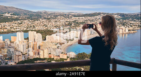 Jeune femme blonde est sur zone clôturée grimpèrent sur Penon de Ifach de Calpe symbole espagnol ville touristique, détient des décors photo prend smartphone Banque D'Images