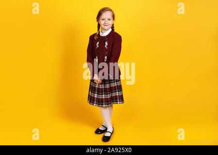 Une écolière est pleine longueur et regarde la caméra. Cute little girl en uniforme sur un fond jaune. Banque D'Images