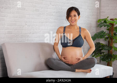 Jeune femme enceinte s'attendant nouvel enfant est assis sur un canapé à la maison. Elle porte des vêtements de sport et à la recherche à l'appareil photo. Banque D'Images