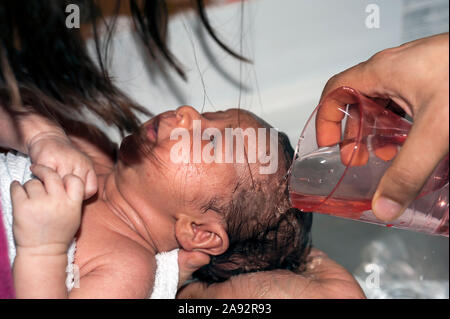 Le verre avec de l'eau versée sur la tête de bébé de deux semaines à pleurer pour laver ses cheveux. Tenu par la mère et bébé lavé par le père's hand holding glass. Banque D'Images