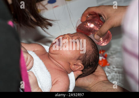 Le verre avec de l'eau versée sur la tête de bébé de deux semaines à pleurer pour laver ses cheveux. Tenu par la mère et bébé lavé par le père's hand holding glass. Banque D'Images