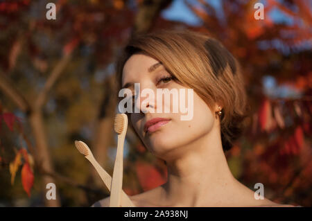 Jeune fille avec de courts cheveux blonds sont titulaires d'une des brosses à dents en bambou à l'extérieur par temps ensoleillé à l'automne sur fond de feuilles rouge et orange Banque D'Images