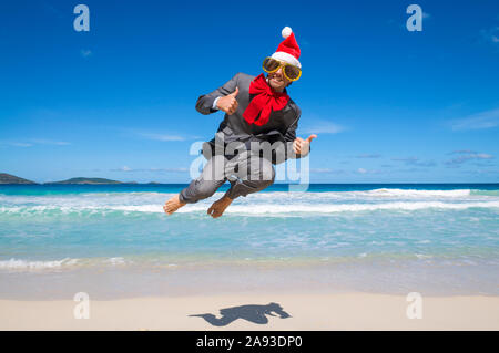 Excited businessman in Santa hat, lunettes et big red Christmas bow sautant sur le rivage d'une plage tropicale Banque D'Images