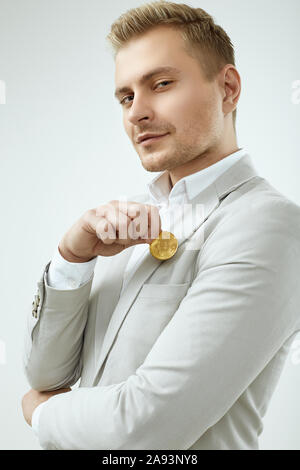 Portrait d'un bel homme blond modèle dans un costume gris de la mode est titulaire d'un Bitcoin isolé sur fond blanc en studio Banque D'Images