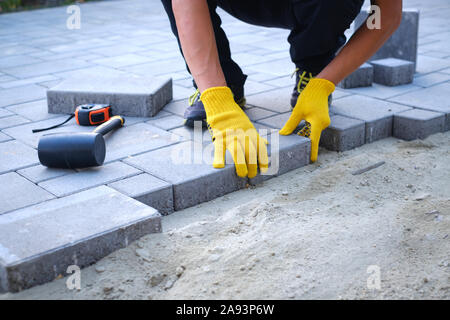 Le master en gants jaunes jette des pavés en couches. Brique de jardin pavage pavé la voie professionnelle par travailleur. La pose des dalles de béton gris dans la chambre cour sur la base de la fondation de sable. Banque D'Images