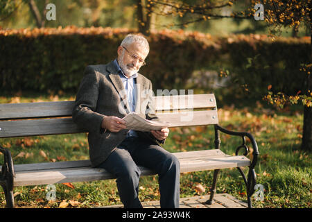 Beau grand-père avec une belle barbe dans un manteau gris s'assied sur un banc dans le parc et lit un journal. Hauts homme aux cheveux gris dans les verres Banque D'Images