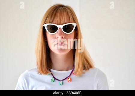 Portrait jeune femme blonde avec expression neutre wearing white cat-eye lunettes de soleil et un puka collier de Banque D'Images