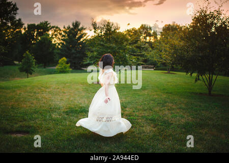 Une femme marche dans la longue robe blanche à travers parc bordé d'arbres au coucher du soleil Banque D'Images