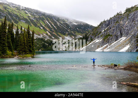 Une femme sort sur le côté d'un lac alpin avec son chien et prend dans la vue dans la chaîne côtière autour de Pemberton (Colombie-Britannique), sur une journée d'été pluvieux. Banque D'Images