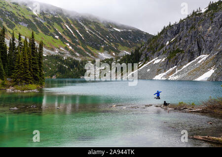 Une femme sort sur le côté d'un lac alpin avec son chien et prend dans la vue dans la chaîne côtière autour de Pemberton (Colombie-Britannique), sur une journée d'été pluvieux. Banque D'Images