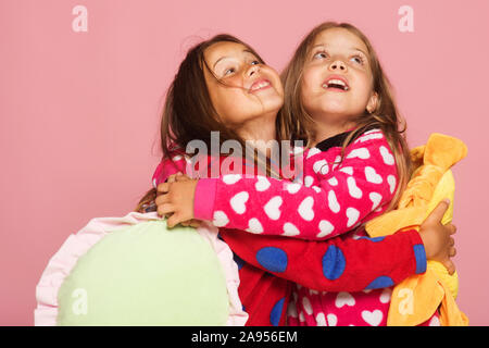 Les enfants ayant surpris les visages et les cheveux hug. Rechercher des enfants et se tenir sur fond rose, copiez l'espace. L'enfance et de bonheur concept. In colorful polka dotted pyjamas tenir oreillers lumineux Banque D'Images