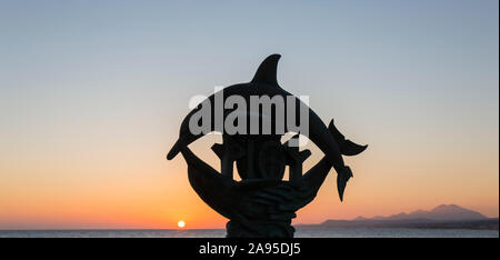 Rethymno, Crète, Grèce. Silhouette de la célèbre statue de dauphin au lever du soleil, Mont Psiloritis visible en arrière-plan. Banque D'Images