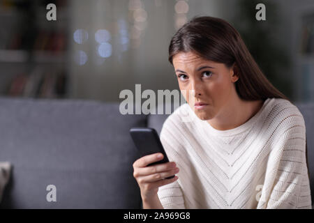 Femme douteuse holding phone caméra regarde assis sur un canapé dans la nuit à la maison Banque D'Images
