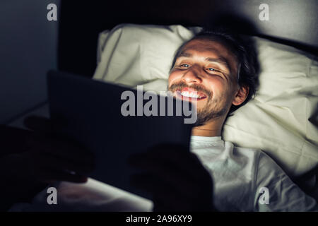 Handsome smiling caucasian male barbu au lit la nuit tout en regardant quelque chose sur sa tablette. Banque D'Images