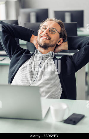 Jeune homme essaie de se détendre tout en écoutant de la musique au travail. Tasse à café et un ordinateur visible au premier plan. Banque D'Images