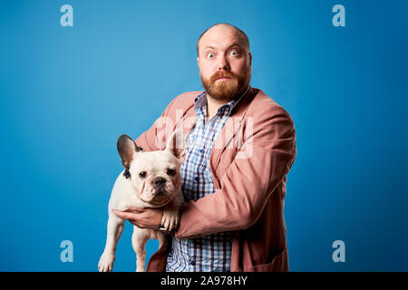 Homme barbu en veste avec bouledogue français dans ses bras sur fond bleu vide en studio Banque D'Images
