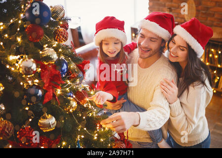 Little girl in Santa hat aidant les parents gais décoration arbre de Noël à la maison Banque D'Images