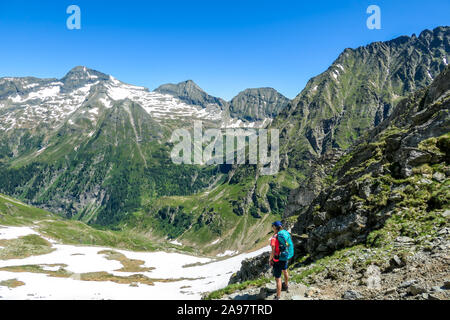 Une jeune femme avec un gros sac à dos de randonnées vers le bas sur un sentier raide entre de hauts sommets de montagnes. Certaines des pentes sont couvertes de neige. Dans le dos est Banque D'Images
