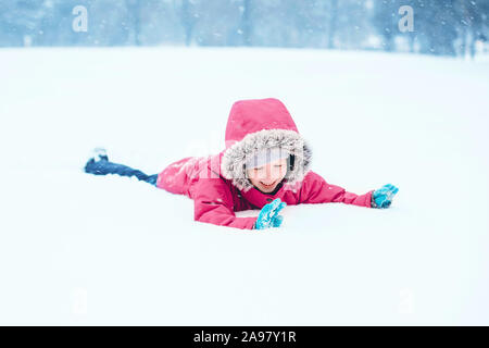 Funny Cute smiling Caucasian girl excité dans l'enfant des vêtements chauds dossier rose jouant avec la neige étendue sur le sol pendant l'hiver froid jour de neige à neige Banque D'Images