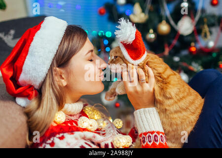 Célébration de Noël avec cat. Femme jouant de l'embrasser et animaux de compagnie à Santa's hat par Nouvel An des arbres à la maison. Banque D'Images