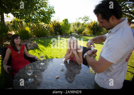 Deux mid-adult woman assis autour d'une table à l'extérieur en attente d'un homme d'ouvrir une bouteille de vin. Banque D'Images