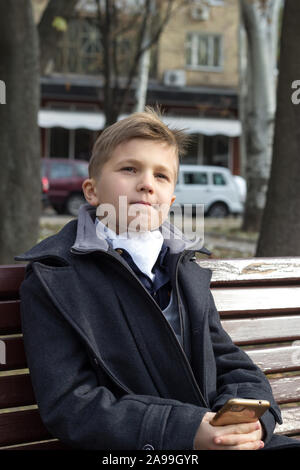 Un garçon est assis sur un banc dans un parc en vêtements d'affaires avec une évaluation de l'aspect et un léger sourire. La réflexion sur son visage, on croise les doigts à la main Banque D'Images