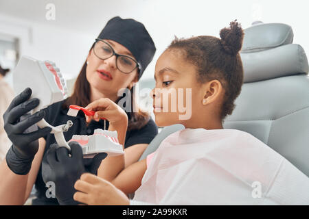 Femme dentiste en uniforme médical et chapeau noir, des lunettes et des gants en caoutchouc modèle dents holding. Enseignement médecin peu patient comment brosser les dents. African girl sitting and holding toothbrush. Banque D'Images