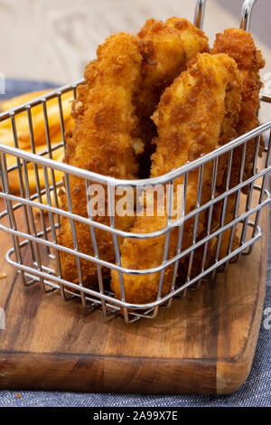 Coujons de poulet enrobées de chapelure dans un fil panier métal, avec des frites, sur une planche à découper en bois Banque D'Images