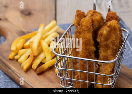 Coujons de poulet enrobées de chapelure dans un fil panier métal, avec des frites, sur une planche à découper en bois Banque D'Images